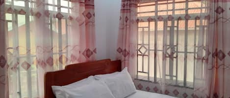 A neatly made bed with white linens and a red blanket, a wooden headboard, and pink patterned curtains with diamond shapes.