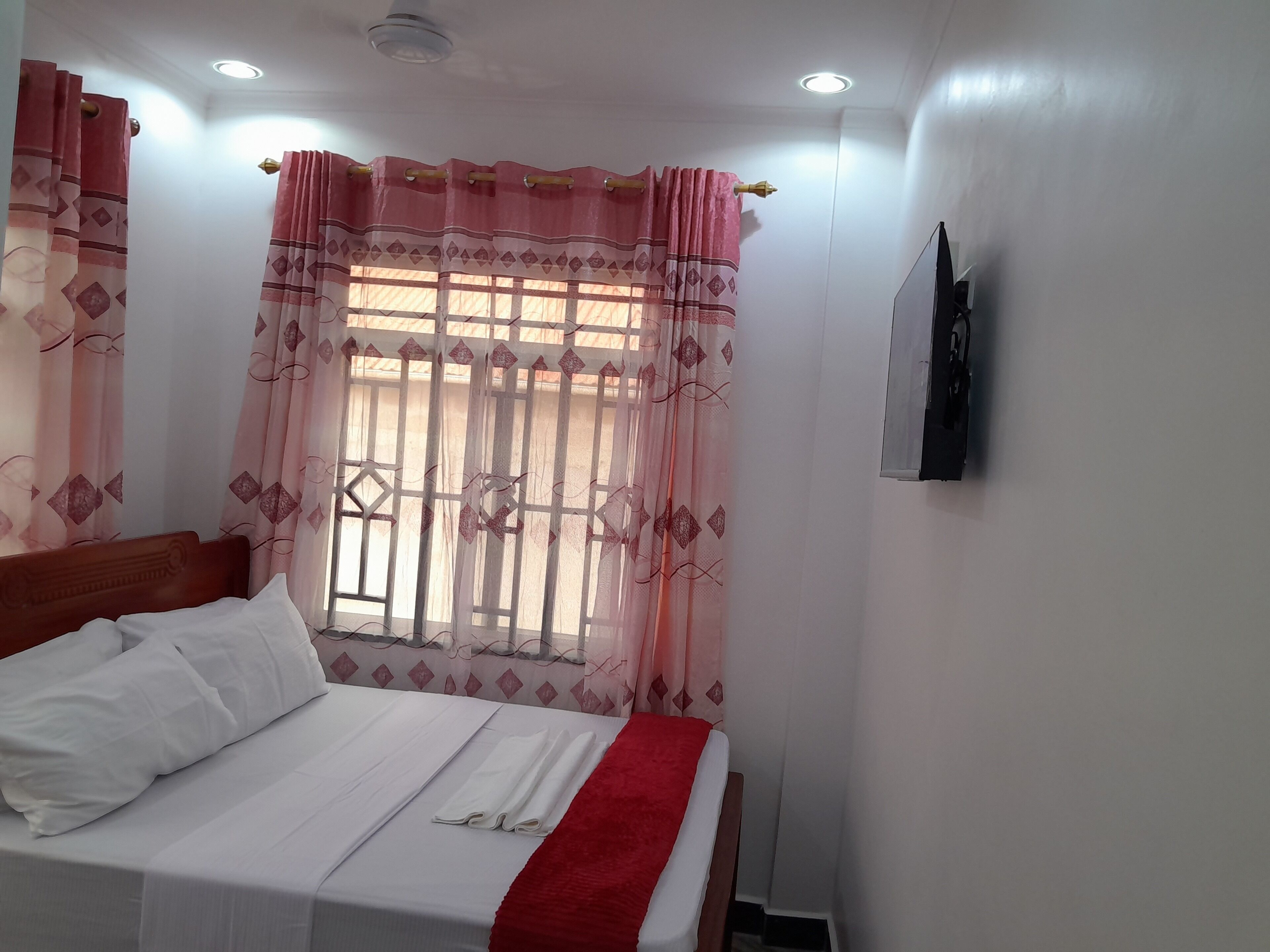 A neatly made bed with white linens and a red blanket, a window with decorative curtains, and a wall-mounted television.