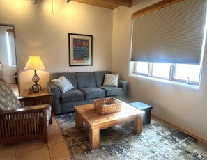 A living room with a sofa, a wooden coffee table, a lamp, and a framed picture on the wall. - Casita Vista Grande: Cozy Adobe with Epic Mountain Views! New on VRBO! Hot Tub (El Prado)
