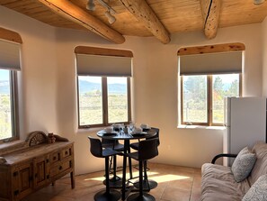 A room with a wooden ceiling, two windows with blinds, a dining table with chairs, a sofa, and a refrigerator.