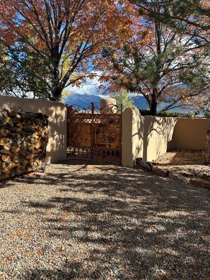 A gated entrance with a pile of firewood, a decorative metal gate, and a gravel driveway leading to a building. - Casita Vista Grande: Cozy Adobe with Epic Mountain Views! New on VRBO! Hot Tub (El Prado)