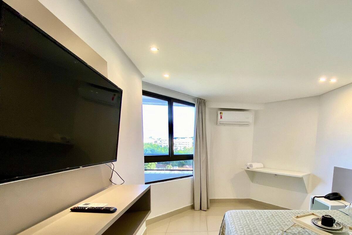 A modern hotel room with a flat-screen TV, a bed with a patterned bedspread, a bedside table with a remote control, and a window with a view of buildings.