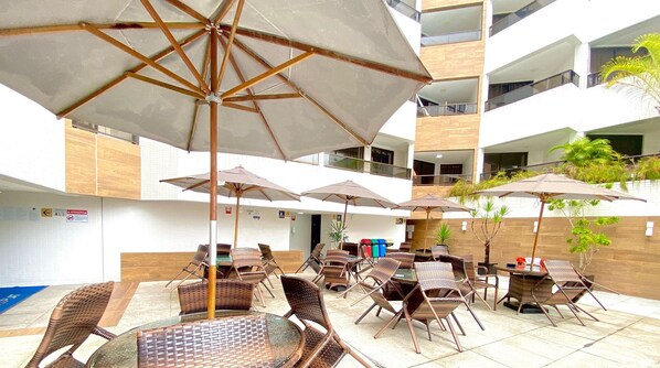 An outdoor patio with wicker furniture and umbrellas, surrounded by a white building with balconies. - Gold Flat Cabo Branco by 123Quarto Hospedagem (João Pessoa)