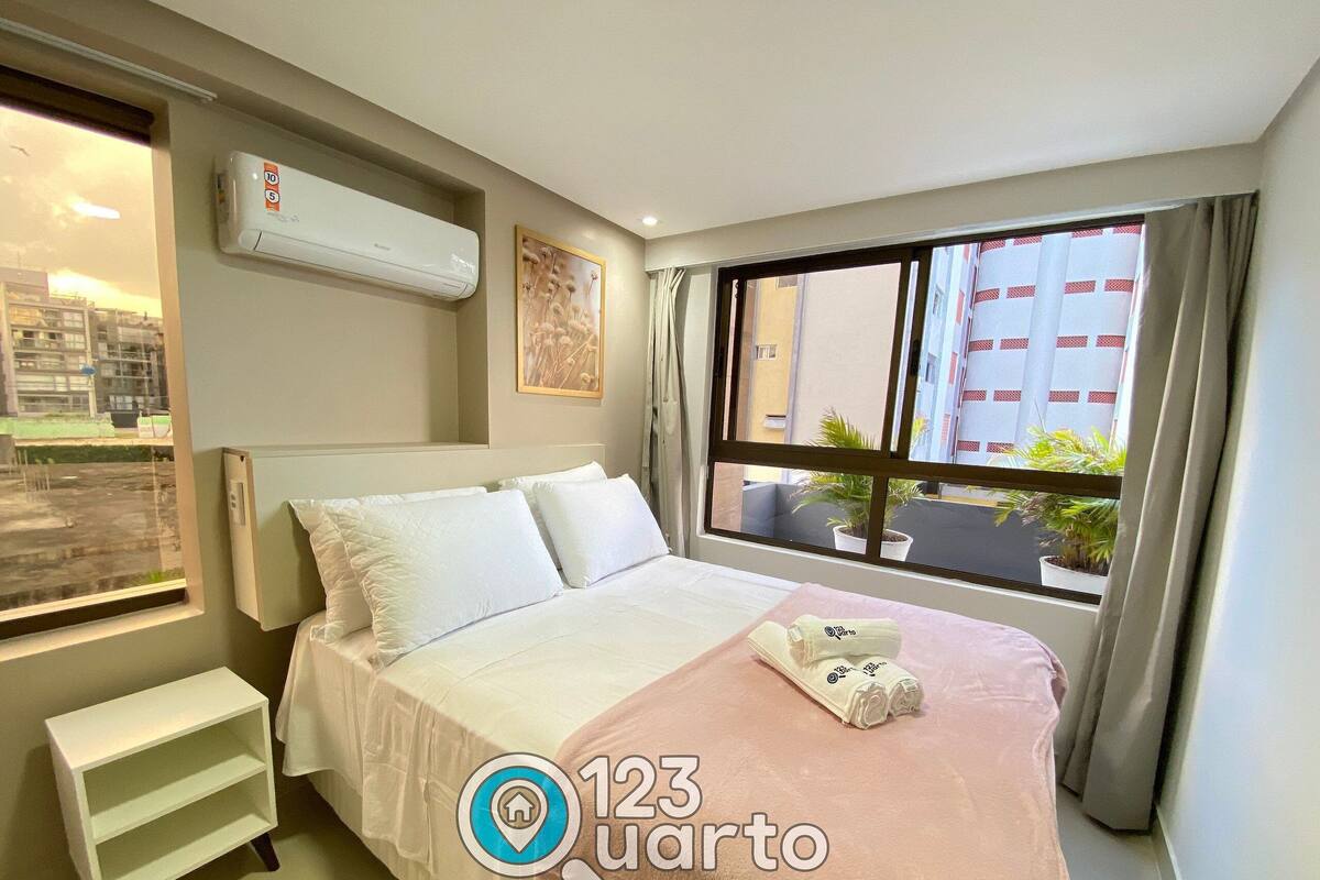 A hotel room with a bed, a small white shelf, an air conditioning unit, a window with a view of buildings, and a framed picture on the wall.