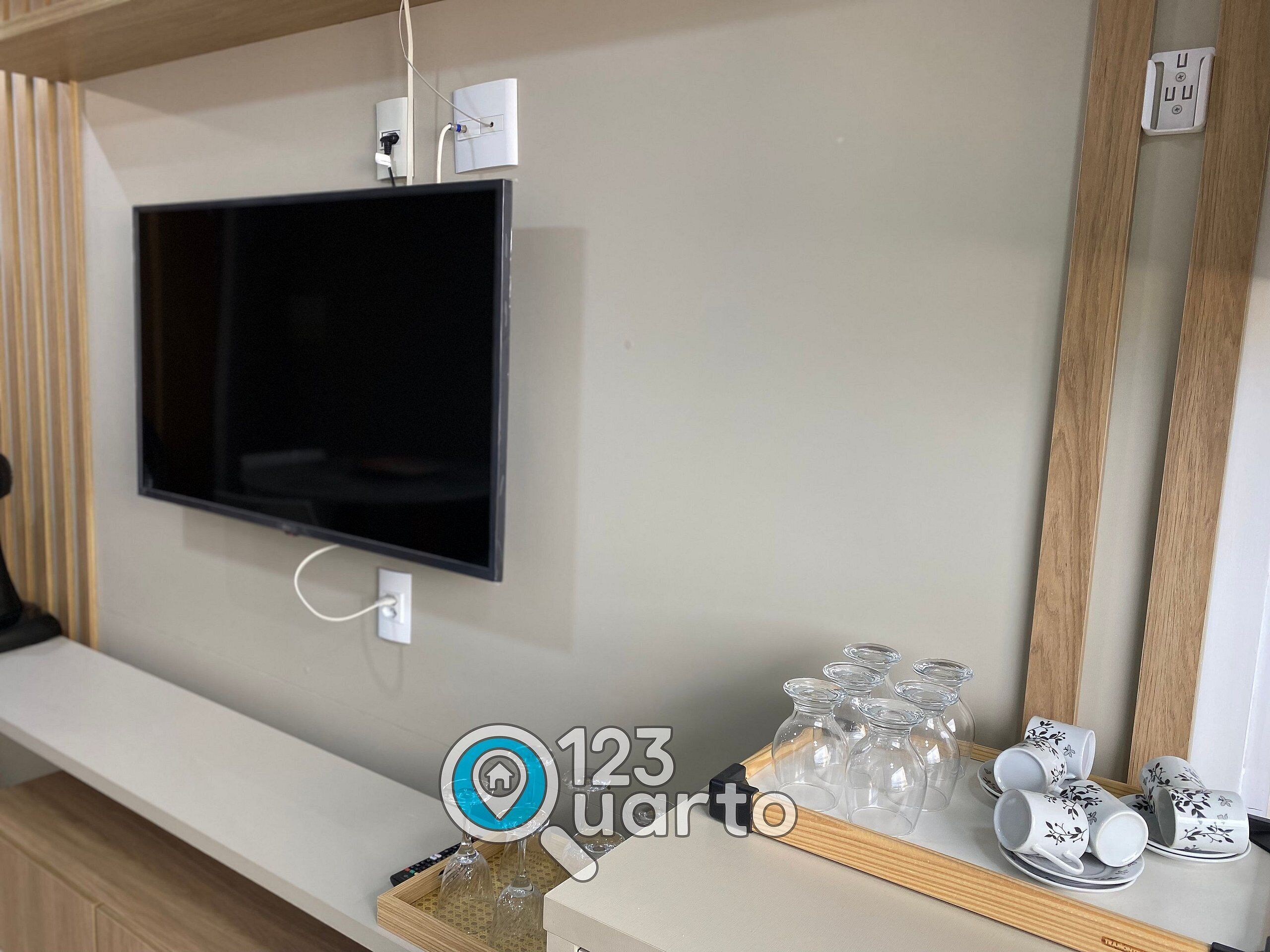 Quarto moderno com televisão na parede, prateleira de madeira com objetos decorativos e espelho.
