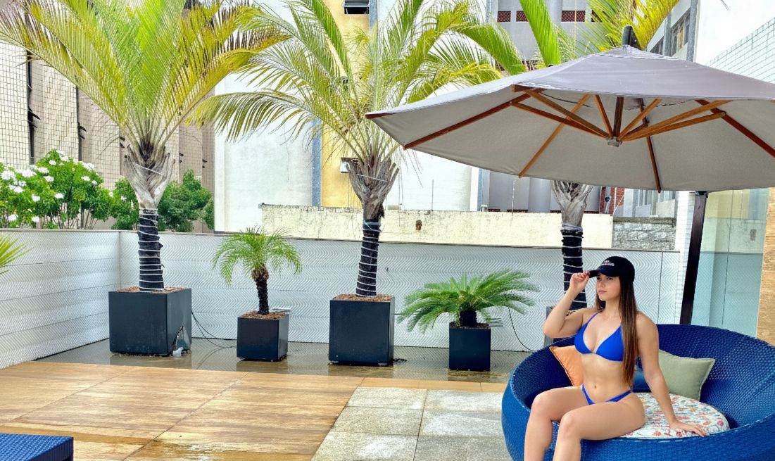 Una mujer sentada en un sillón de bolas azules debajo de un paraguas grande en un patio al aire libre con palmeras y plantas en macetas.