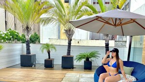 A woman sitting on a blue bean bag chair under a large umbrella in an outdoor patio area with palm trees and potted plants. - Gold Flat Cabo Branco by 123Quarto Hospedagem (João Pessoa)