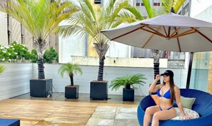 Una mujer sentada en un sillón de bolas azules debajo de un paraguas grande en un patio al aire libre con palmeras y plantas en macetas.