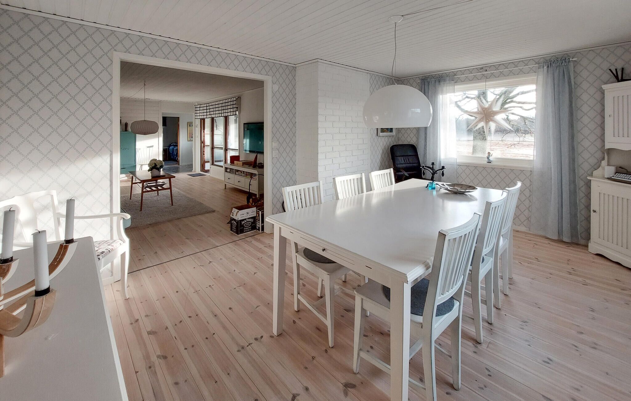 Un vaste espace dédié à la salle à manger, avec une table et des chaises blanches, une cuisine en arrière-plan et une grande fenêtre offrant une vue dégagée.