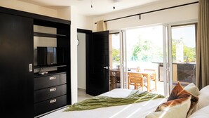Ein modernes Hotelzimmer mit einem großen Bett, einem Fernseher auf einer schwarzen Kommode, einem Balkon mit Tisch und Stühlen sowie einer Schiebeglastür nach draußen.