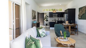 Ein modernes Wohnzimmer mit einer Couch, grünen Kissen, einem Couchtisch mit einer Vase und Blick in den Küchenbereich mit dunklen Schränken und einem Esstisch.
