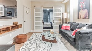 A modern living room with a glass coffee table, a sofa with cushions, a television mounted on the wall, and a sliding door leading to a bedroom. - Pet-Friendly Apt  Fenced yard  Private Parking (Allentown)
