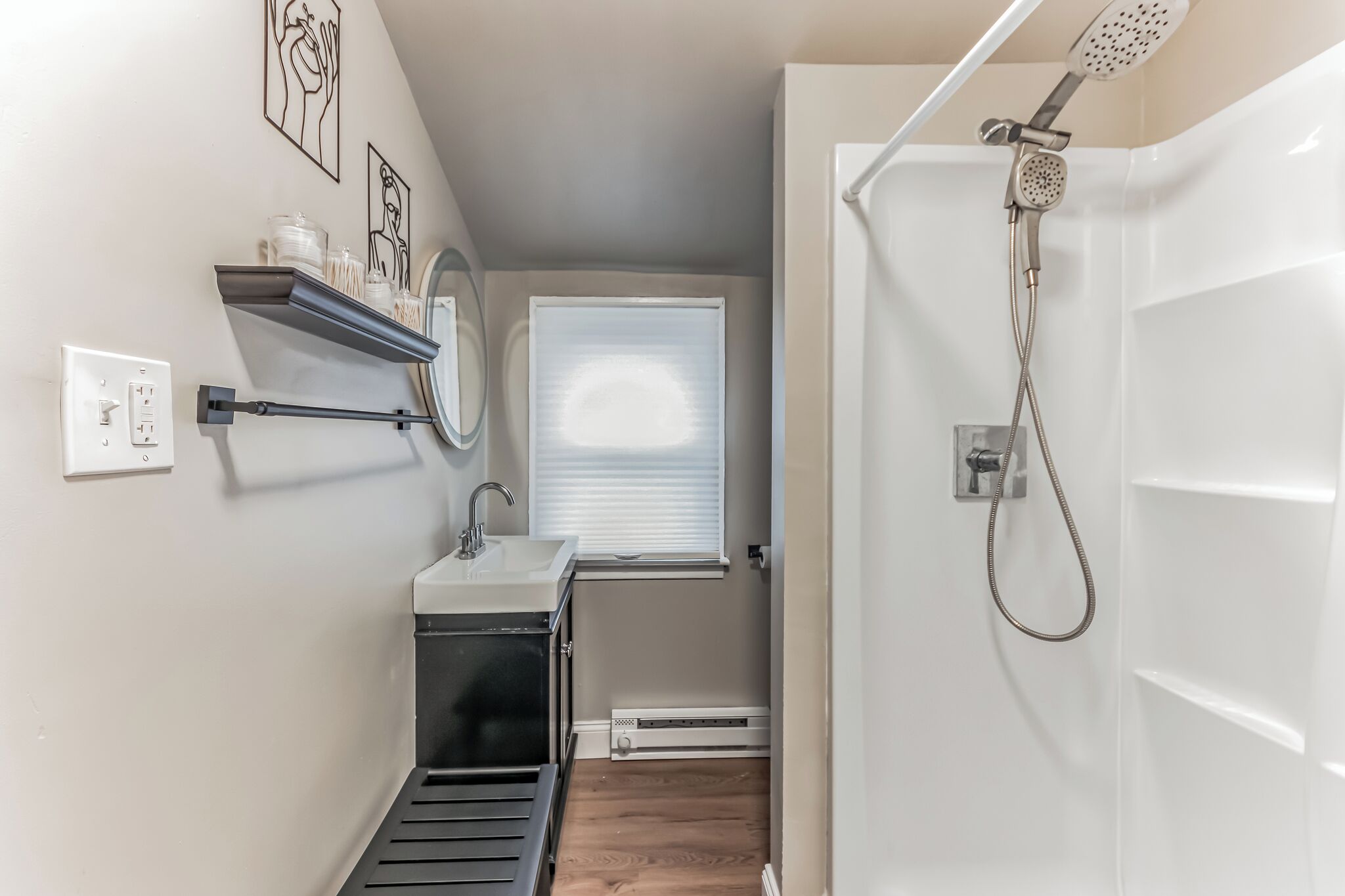 A compact bathroom with a sink, mirror, shower, and a window with blinds.