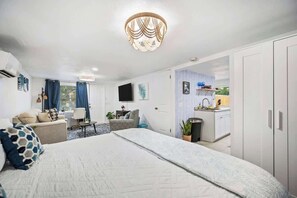 A hotel room with a large bed, a desk, a chair, a TV, a kitchenette, and a wardrobe.