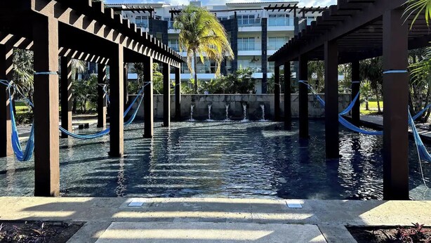 Zona de piscina con pérgolas de madera y hamacas, rodeada de palmeras y un edificio al fondo.