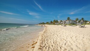 Una playa con orilla arenosa, olas del océano y un edificio con varios balcones y palmeras al fondo.