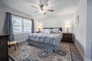 Une chambre à coucher avec un lit, deux tables de chevet avec des lampes, un ventilateur de plafond, une fenêtre avec des rideaux et un tapis à motifs.