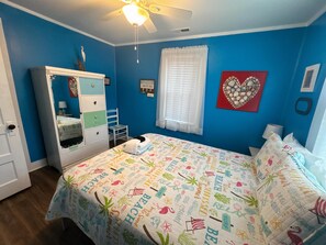 Une chambre à coucher comprenant un lit, une commode, une fenêtre avec des stores, un ventilateur de plafond et un mur orné d’un décor en forme de cœur.