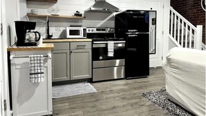 Een compacte keuken met een magnetron, fornuis, oven en een zwarte koelkast. Boven het aanrecht zijn houten planken en er is een witte deur op de achtergrond te zien.