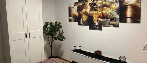 Une chambre à coucher avec un lit, une armoire blanche, une plante en pot et une étagère murale agrémentée d’objets décoratifs.