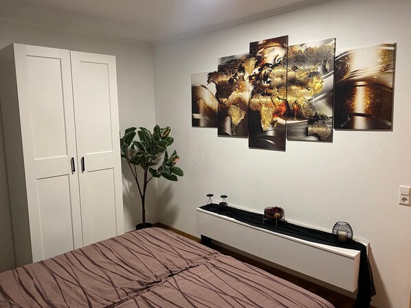 A bedroom with a bed, a white wardrobe, a potted plant, and a wall-mounted shelf with decorative items. - Vacation apartment Delja (Aulendorf)