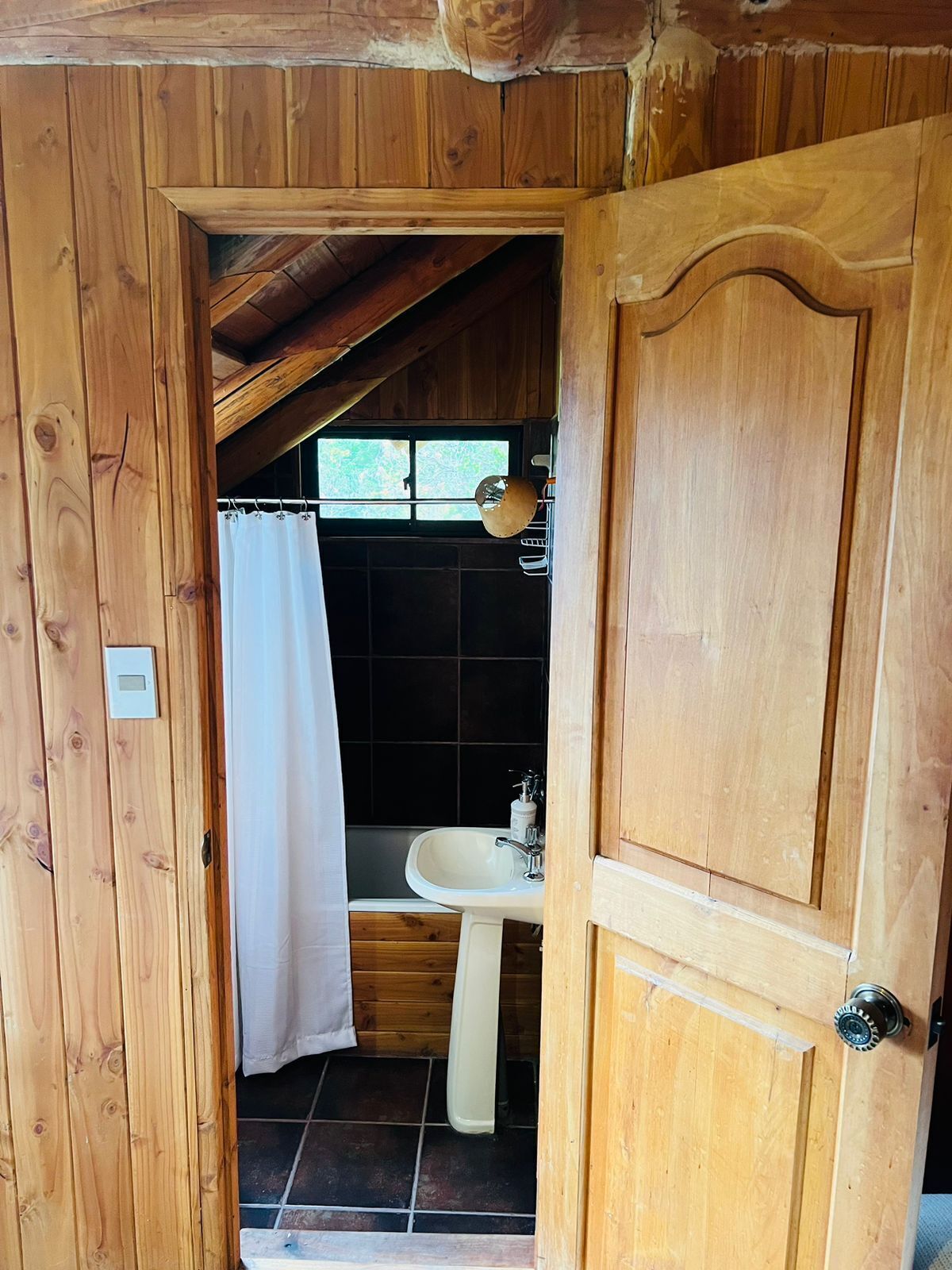 Une salle de bain rustique, avec des murs en bois, un lavabo blanc, un rideau de douche et une fenêtre au-dessus du lavabo.
