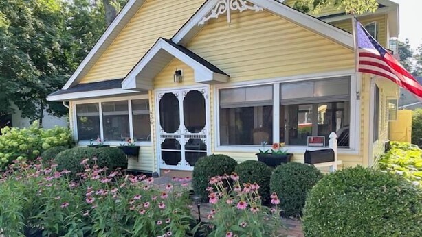 A yellow house with a porch, a flag, and a garden with pink flowers.
