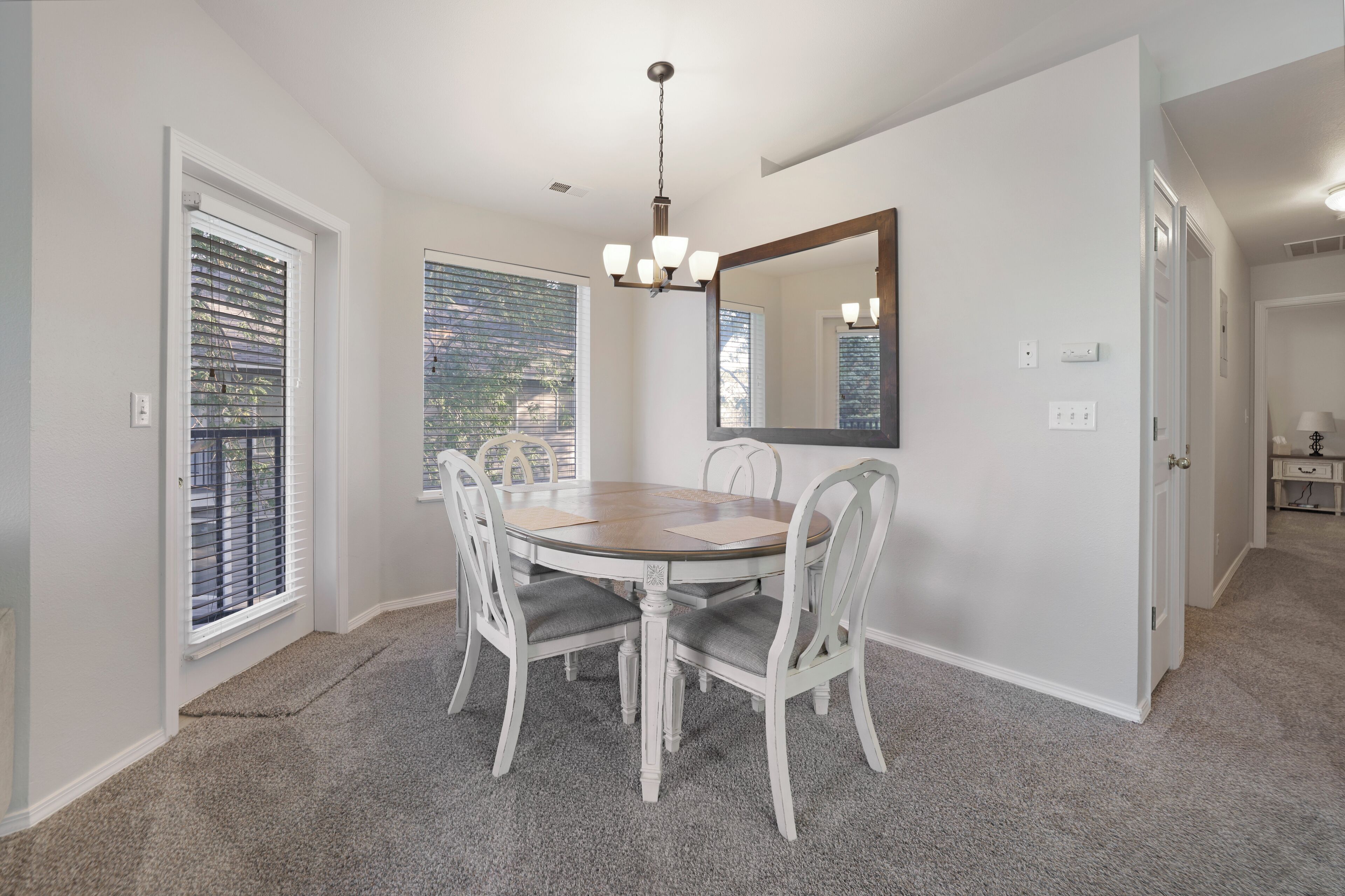 A dining area with a round table, white chairs, a chandelier, and a large mirror on the wall.