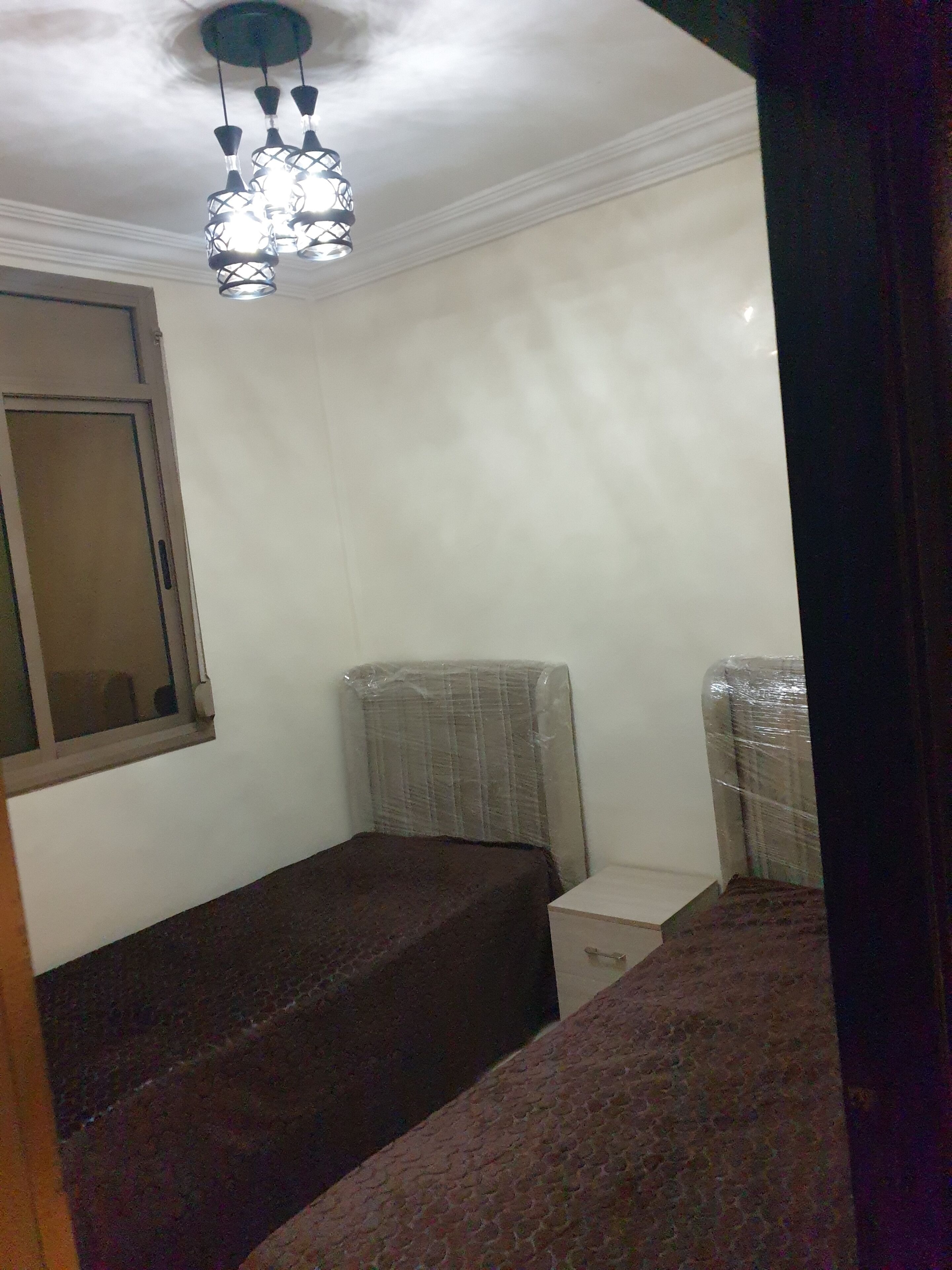 Ein kleiner, schwach beleuchteter Raum mit zwei Betten, einem Fenster mit Holzrahmen und einer Deckenleuchte.