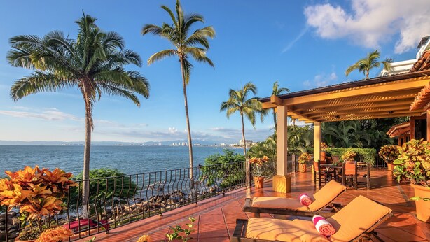 Una terraza con sillones de sol, barandilla y vista al mar y palmeras.
