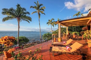 A terrace with sun loungers, a railing, and a view of the sea and palm trees.