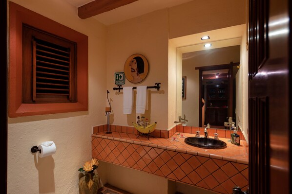 A bathroom with a toilet paper holder, a towel rack with two towels, a sink with a mirror, and a window with blinds.
