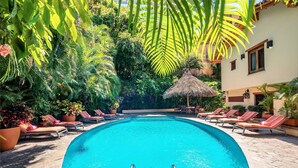Una piscina rodeada de sillas de descanso y plantas en macetas, con una estructura de techo de paja al fondo.