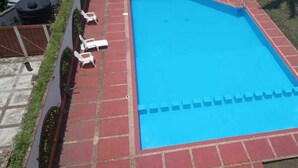 Una piscina rectangular con revestimiento azul, rodeada por una terraza de baldosas rojas. Se observan sillones de descanso blancos alineados a lo largo del borde de la piscina.