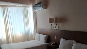Habitación de hotel con dos camas, una mesita de noche, una lámpara, un equipo de aire acondicionado y una ventana con cortinas.