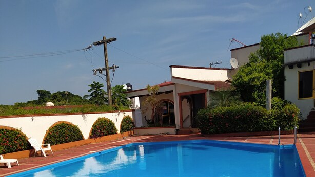 Una piscina con escalones de acceso, rodeada por un jardín con diversas plantas y un edificio blanco de techo rojo.