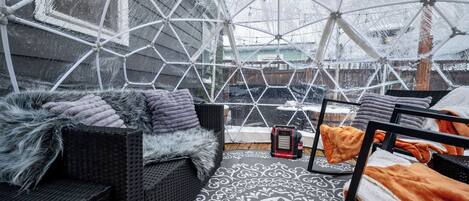 Uma área de estar externa acolhedora e protegida, com teto de vidro geométrico, móveis de vime e tapete estampado.