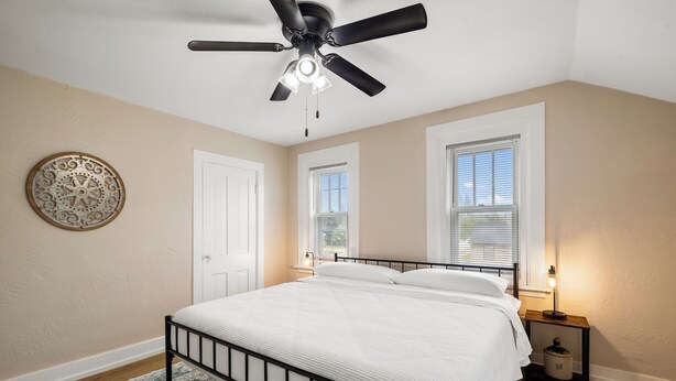 Un dormitorio con cama, ventilador de techo, dos ventanas con persianas, una mesita de noche con lámpara y un adorno en la pared.