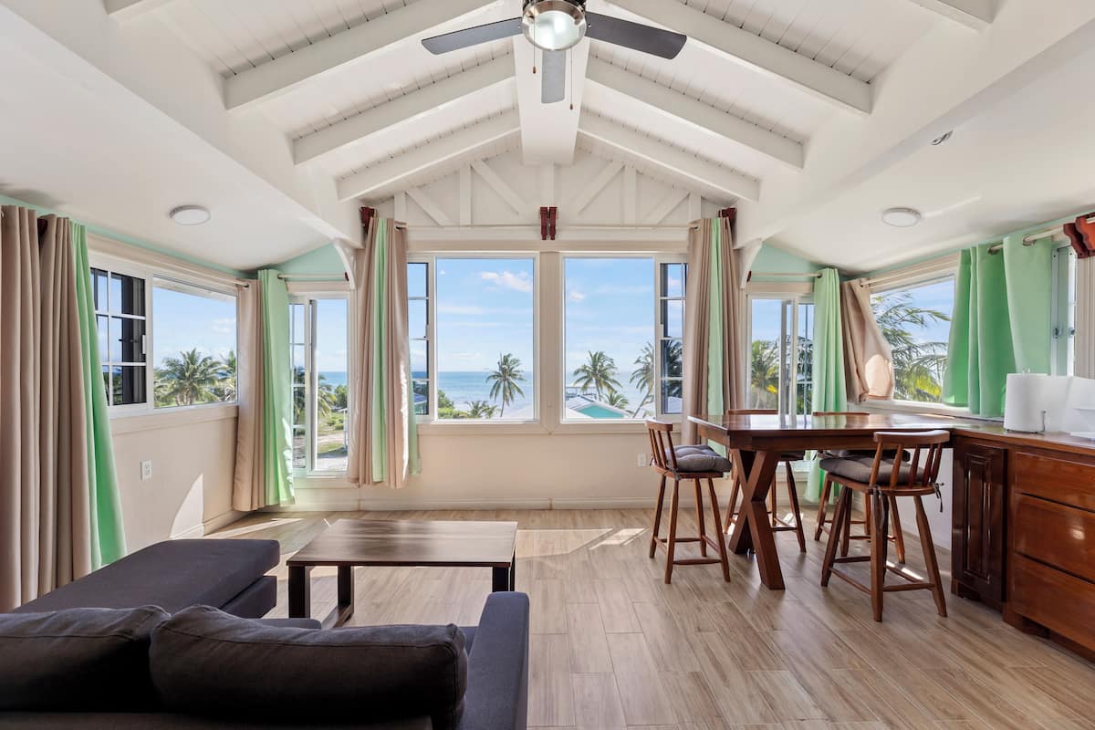 Una habitación amplia con techo de madera, ventanas grandes que dan al océano, una zona de comedor con mesa y sillas de madera, y un sofá.