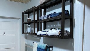 Un baño con una puerta blanca, un perchero con una toalla azul, y un estante con toallas dobladas y artículos de aseo.