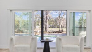 Een moderne kamer met twee witte fauteuils, een klein rond tafeltje en uitzicht op een park door grote glazen deuren.
