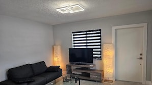A living room with a black sofa, a glass coffee table, a flat-screen TV on a wooden stand, and two wall-mounted lamps. - Charming 2-bedroom apartment in terrific Hollywood with AC, WiFi (Hollywood)