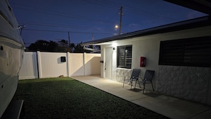 A residential backyard with a lawn, a patio with two chairs, a white fence, and a house with a stone accent wall. - Charming 2-bedroom apartment in terrific Hollywood with AC, WiFi (Hollywood)
