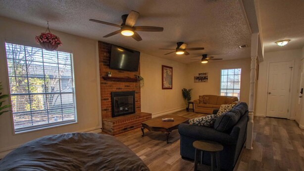 A living room with a fireplace, ceiling fans, a sofa, and a large window. - Spacious Stay for 16 - Central Fort Worth (Fort Worth)