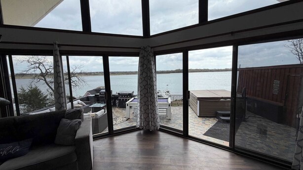 A spacious room with large windows overlooking a lake, featuring a wooden floor, a sofa, and outdoor grilling equipment.