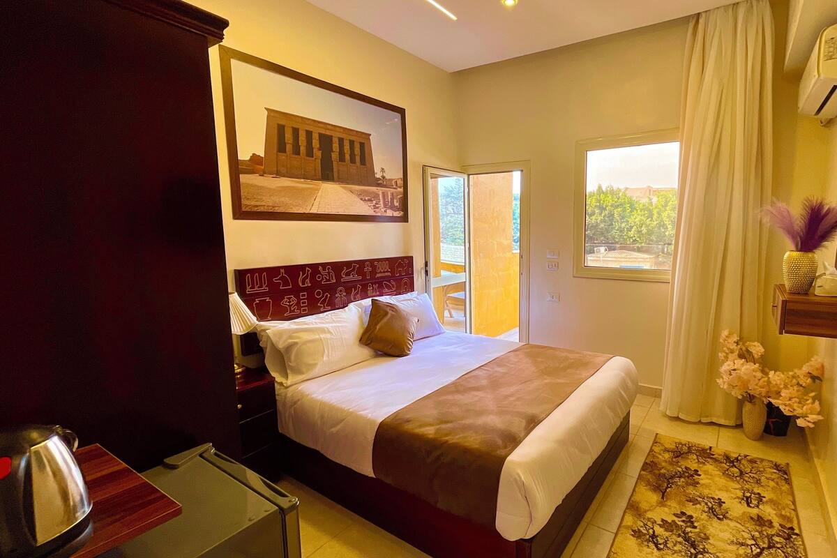 A hotel room with a bed, a flat-screen TV, a window with curtains, a small table with a lamp, and a decorative wall piece.