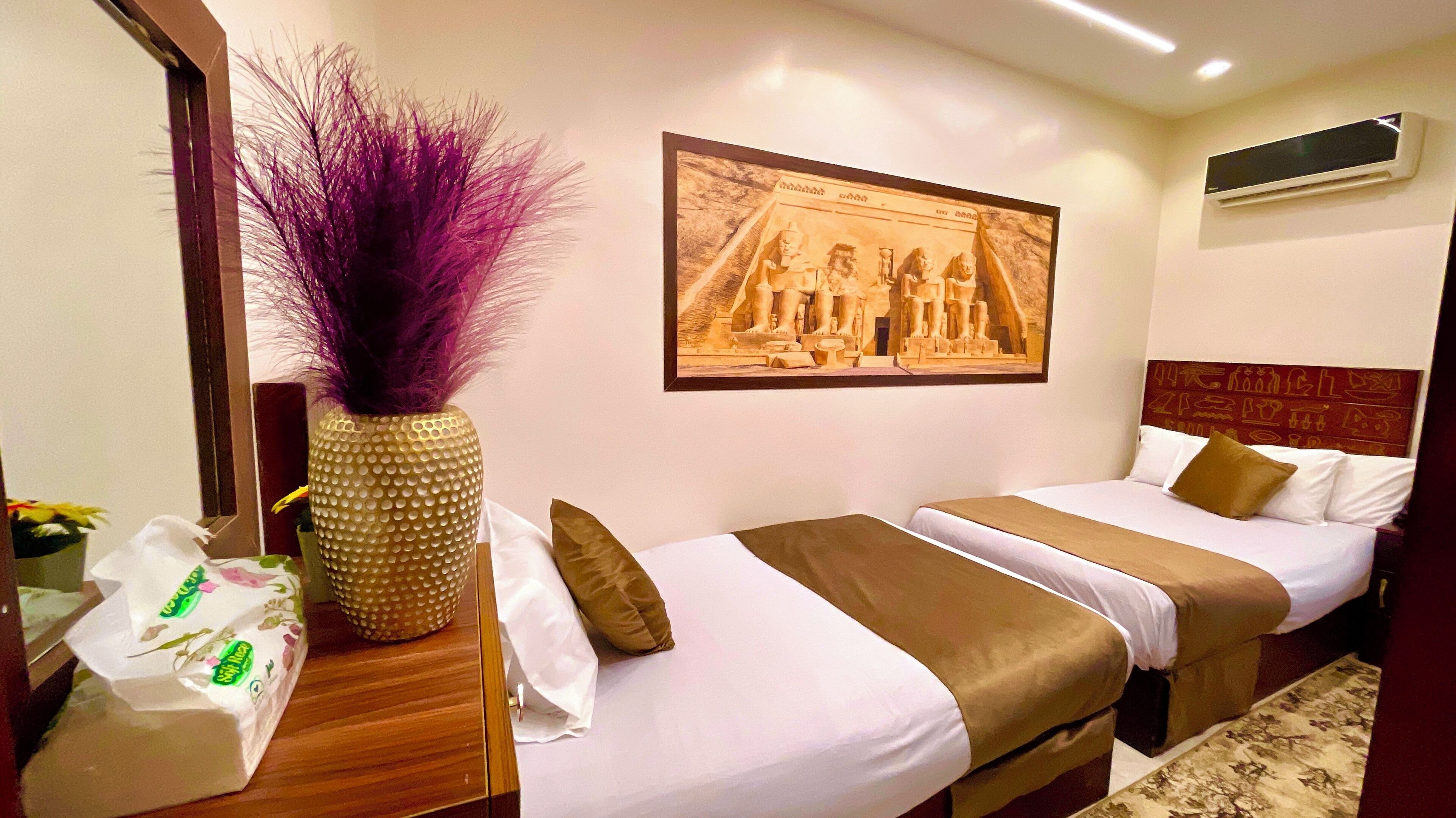 A hotel room with two beds, a large mirror, a framed picture on the wall, and a vase with purple flowers.