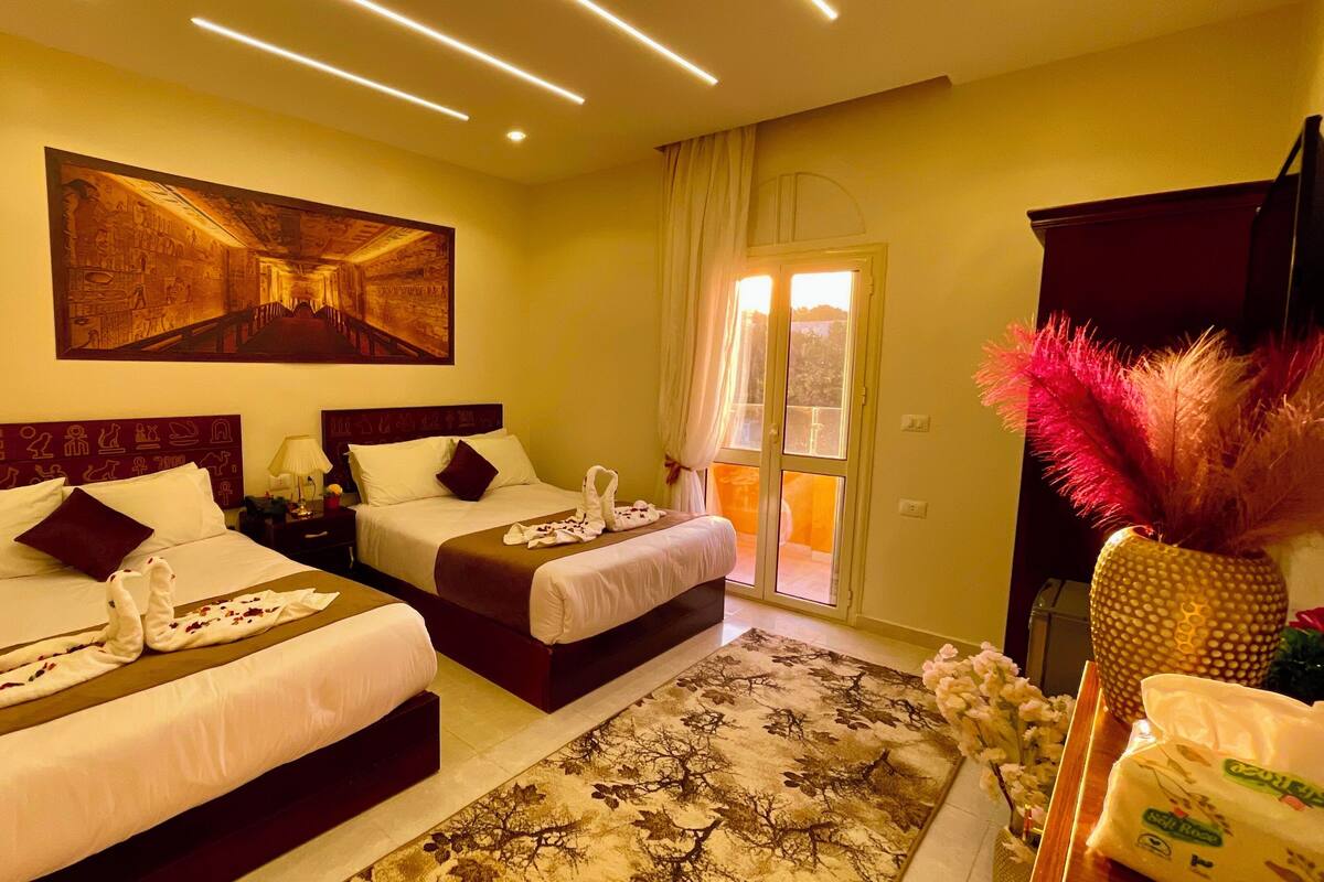 A hotel room with two beds, a large painting on the wall, a view of the outdoors through a sliding door, and a decorative vase with pink feathers.