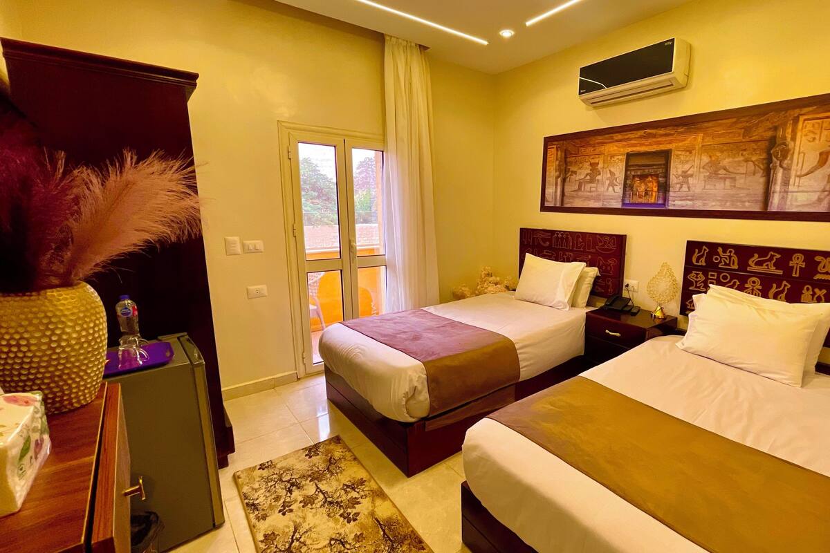A hotel room with two beds, a large window with curtains, an air conditioning unit, and a wall with Egyptian-themed artwork.