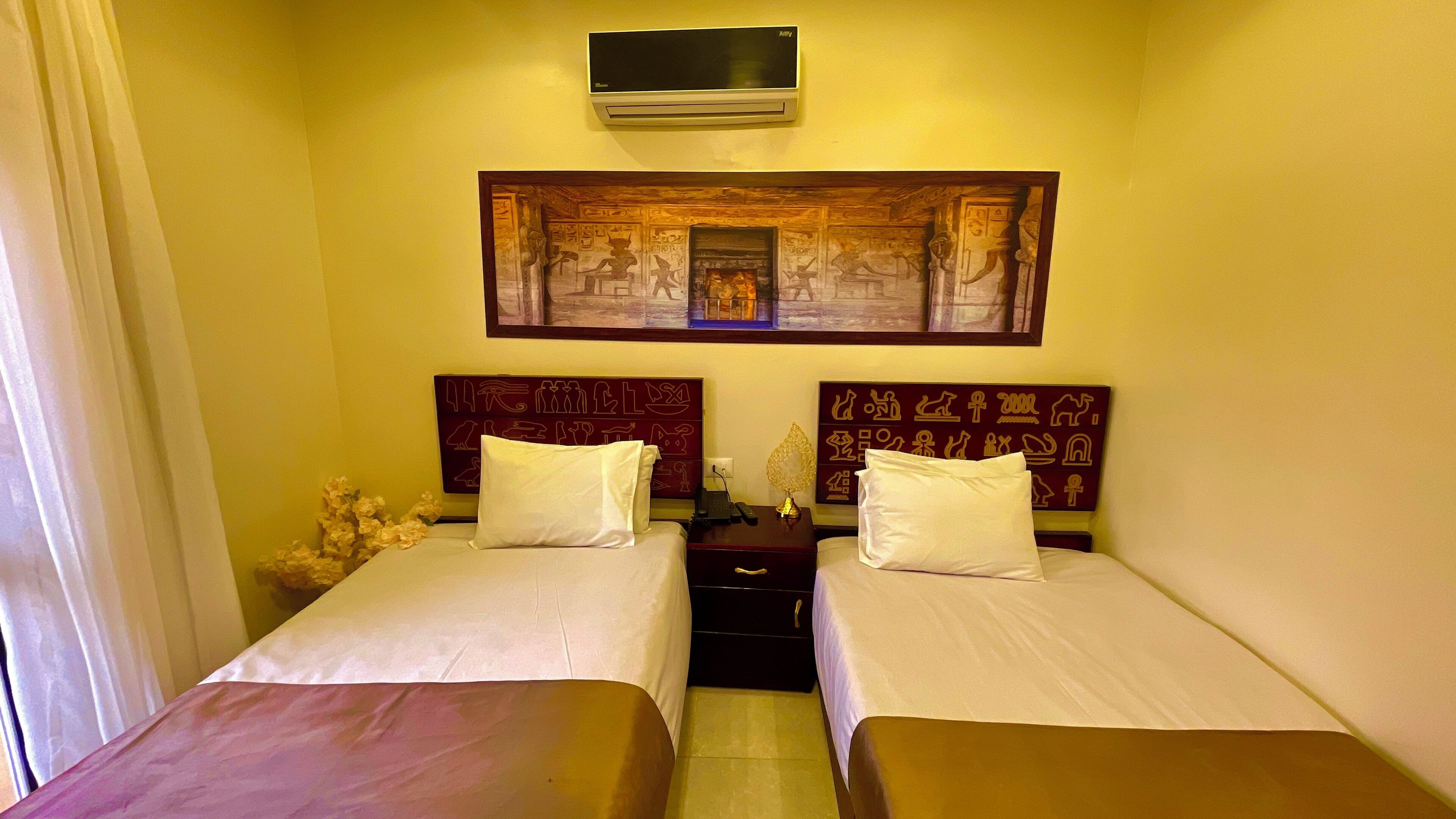 Quarto de hotel com duas camas, uma mesa de cabeceira, aparelho de ar condicionado e uma parede com arte decorada ao tema egípcio.